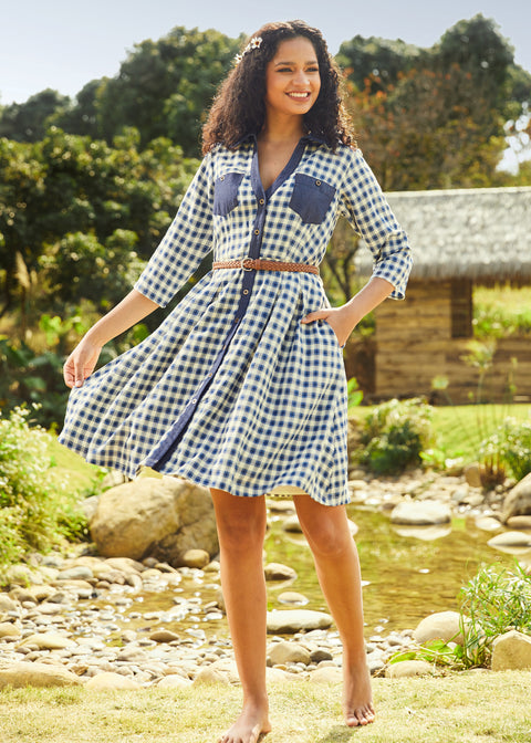 A woman standing outdoors in a casual chic setting, wearing a blue and white checkered shirtdress with long sleeves, a round neckline, and a belt.